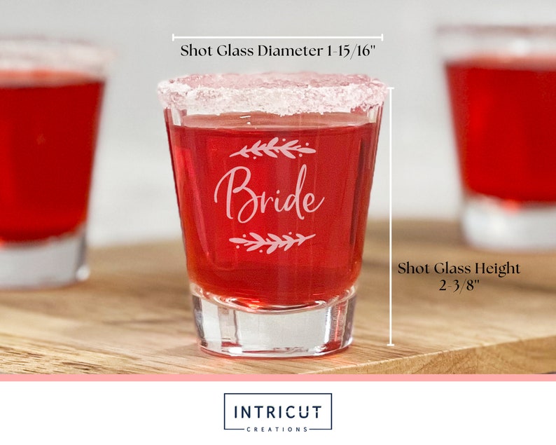 Puede incluir: Un vaso de chupito transparente lleno de l&iacute;quido rojo, con borde blanco y grabado con la palabra "Bride" y detalles florales. El vaso de chupito mide 6 cm de alto y 5 cm de di&aacute;metro. El vaso de chupito est&aacute; sobre una superficie de madera.