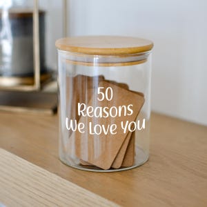May include: A clear glass jar with a wooden lid, filled with small brown cards. The jar has white text that reads "50 Reasons We Love You". The jar is sitting on a wooden surface.