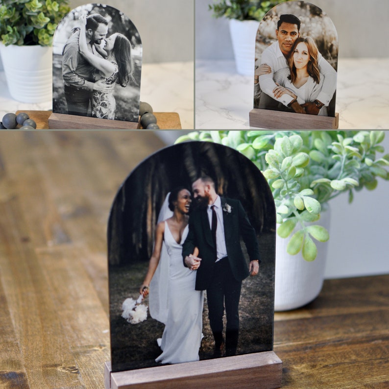 Puede incluir: Un conjunto de tres expositores de fotos con forma de arco con bases de madera. Cada expositor presenta una pareja diferente en una fotograf&iacute;a. La primera foto muestra una pareja bes&aacute;ndose, la segunda muestra una pareja abraz&aacute;ndose y la tercera muestra una pareja caminando por un bosque.