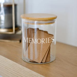 May include: A clear glass jar with a wooden lid, filled with brown cards. The word "MEMORIES" is printed in white on the front of the jar. The jar is sitting on a light-colored wooden surface.