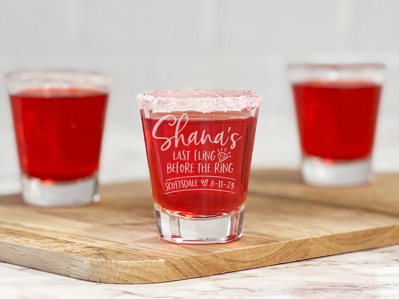 Puede incluir: Tres vasos de chupito llenos de l&iacute;quido rojo, uno enfocado con el texto "Shana's Last Fling Before the Ring Scottsdale 8-11-23". El borde del vaso central est&aacute; recubierto con una sustancia blanca, probablemente sal o az&uacute;car. Los vasos est&aacute;n sobre una tabla de madera.