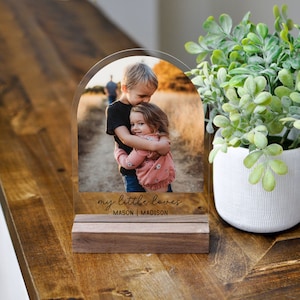 May include: A clear acrylic photo display with an arched top, featuring a color photograph of two children hugging. The base is made of dark wood, and the text "my little loves" and names are printed on the front. A potted plant is in the background.