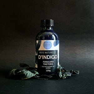 May include: A dark blue bottle with a black cap, labeled "Pâte Naturelle D'Indigo" with text in French. The bottle is surrounded by dried leaves and has a 120g weight listed. The label also includes the text "Dahlia Milon".