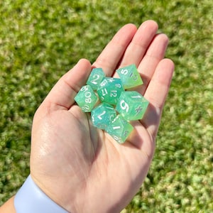 May include: A hand holding a set of seven green and white dice with white numbers. The dice are translucent and have a glittery finish.