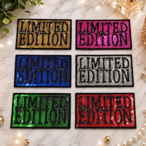 Peut inclure: Six écussons rectangulaires à bordure noire, chacun portant le texte LIMITED EDITION en lettres capitales noires. Le fond de chaque écusson est recouvert de paillettes aux couleurs or, rose, bleu, argent, vert et rouge respectivement.