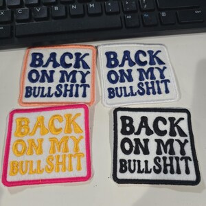 May include: Four square embroidered patches with the text "BACK ON MY BULLSHIT". The patches have different color schemes: orange border with blue text, white with blue text, pink with yellow text, and black with white text.