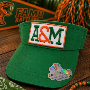 May include: A green visor featuring a rectangular patch with the letters A&M in green and orange on a white background. A holographic sticker on the visor displays the text Creative Love T-shirt & Vinyl Supply Shop and the phone number 469-843-9886.