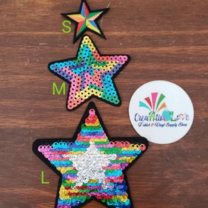 May include: Three rainbow sequin star iron-on patches in different sizes: small, medium, and large. The patches are on a wooden surface.