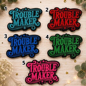 Peut inclure: Cinq écussons brodés numérotés de un à cinq, affichant le texte TROUBLE MAKER dans une police stylisée. Les écussons sont disponibles en bleu sarcelle, rouge, bleu, vert et rose, sur un fond noir pailleté avec des volutes et des étoiles.