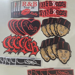 May include: A collection of embroidered patches featuring various designs including R&B Tour 26 with a red lip icon, BREEZY in red text, black heart shapes with R&B, stylized red lettering, rectangular patches with the text USHER, and portraits of a person.