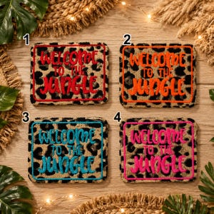 Peut inclure: Quatre dessous de verre rectangulaires avec un motif léopard sur fond beige. Chaque dessous de verre présente le texte brodé "WELCOME TO THE JUNGLE" dans une couleur différente : rouge, orange, turquoise et rose vif, avec une bordure assortie.