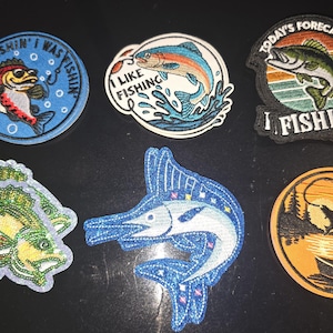 May include: A collection of six embroidered fishing patches. Designs include fish, fishing scenes, and text like "Wishin' I Was Fishin'" and "I Like Fishing". Colors include blue, green, orange, and white.