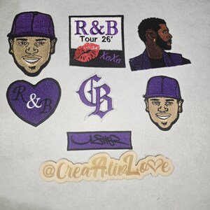 May include: A collection of embroidered patches featuring images of a singer, including a profile view, and text that reads "R&B Tour 26'" with a kiss mark, and the word "Usher". The patches are in purple, tan, and white.