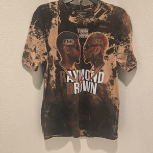 May include: A black and tan acid-wash t-shirt featuring a graphic of two men facing each other with the text TOUR 2026, RNB, and RAYMOND & BROWN printed on the front. The shirt has short sleeves and a crew neckline, displayed on a black plastic hanger.