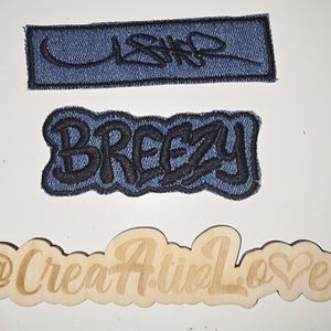May include: Three patches and a wooden cutout are displayed. Two denim patches feature black embroidered text: "USHR" and "BREEZY." A wooden cutout reads "@CreatiA.tinLove" with a heart symbol.