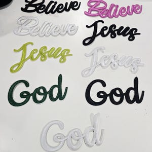 May include: A collection of embroidered fabric patches featuring the words Believe, Jesus, and God in various colors including black, pink, white, lime green, and dark green. Each patch is rendered in a cursive script font with a raised texture.