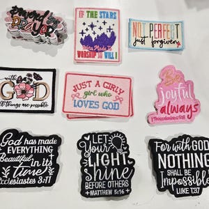 May include: A collection of nine embroidered patches featuring various religious phrases and designs. The patches include text such as Powered by Prayer, If the Stars Were Made to Worship, Not Perfect Just Forgiven, With God All Things Are Possible, Just a Girly Girl Who Loves God, Be Joyful Always, God Has Made Everything Beautiful, Let Your Light Shine, and For With God Nothing Shall Be Impossible.