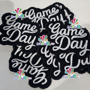 May include: A collection of black embroidered patches with the words "Game Day" in white script. The patches have a decorative border and are arranged in a slightly overlapping pattern.