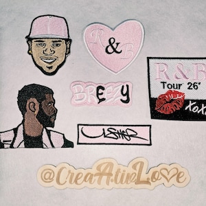 May include: A collection of embroidered patches featuring a portrait of a person in a pink cap, a pink heart with R&B, a BREEZY patch, a rectangular patch with R&B Tour 26' and a red lip print, an USHER patch, and a wooden sign reading @CreaA.tinLove.