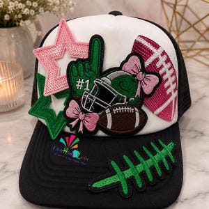 May include: A white and black trucker hat featuring various patches including a green foam finger with #1, a football helmet with a pink bow, a brown football, a pink sequined star, a green star, and a green football lace patch on the brim.