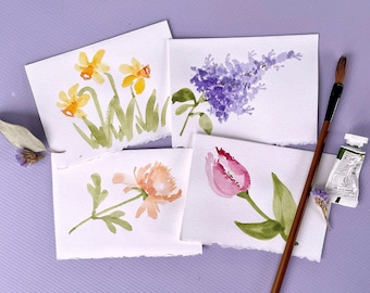 Op de afbeelding: Vier aquarellen van bloemen op wit papier met gescheurde randen. De bloemen zijn narcissen, lila, een enkele tulp en een enkele bloem met oranje bloemblaadjes. Een penseel en een tube verf zijn ook op de afbeelding te zien.