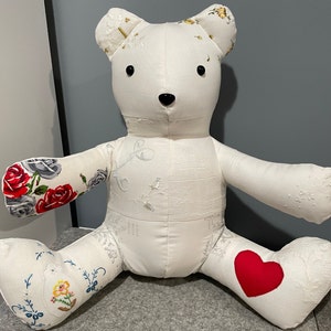 May include: A white patchwork teddy bear with embroidered floral details. The bear has a red heart on one foot and a floral design on the other.