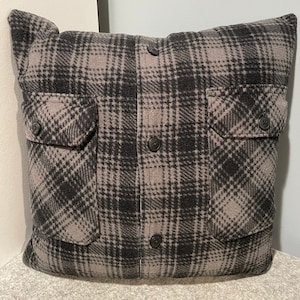 May include: A gray and black plaid pillow with two pockets and buttons. The pillow is made of a soft, fuzzy fabric.