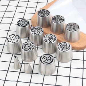 May include: A collection of stainless steel piping tips with various decorative patterns. The tips are arranged on a wooden cutting board and a checkered surface. Two smaller, cone-shaped tips are also included.