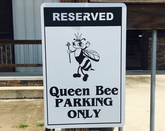 May include: A white sign with black text that reads "Reserved" and "Queen Bee Parking Only". A cartoon bee wearing a crown and holding a scepter is pictured on the sign.