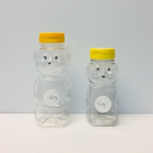 May include: Two clear plastic bear-shaped honey bottles with yellow lids. The larger bottle is labeled "12oz" and the smaller bottle is labeled "8oz".