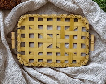 May include: A rectangular bamboo woven tray with wooden handles. The tray has a light brown color and a woven pattern. The handles are made of light-colored wood. The tray is sitting on a wrinkled, light-colored fabric.