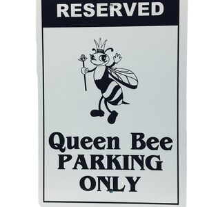 Puede incluir: Un letrero de metal blanco con texto negro y una abeja de dibujos animados con una corona. El letrero dice "RESERVED Queen Bee PARKING ONLY".