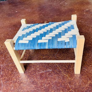 May include: Small wooden stool with a woven seat. The seat features a blue and white pattern. The legs and frame are made of light-colored wood. The stool is a decorative and functional piece of furniture.