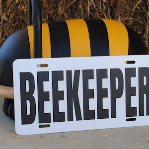 Puede incluir: Un letrero blanco con la palabra "BEEKEEPER" impresa en letras negras. El letrero est&aacute; apoyado en un juguete con forma de abeja negro y amarillo.