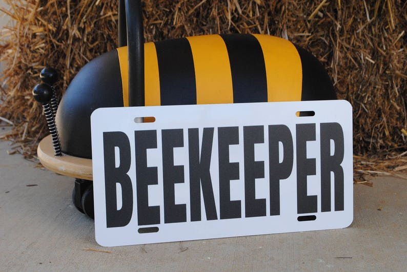 Puede incluir: Un letrero blanco con la palabra "BEEKEEPER" impresa en letras negras. El letrero est&aacute; apoyado en un juguete con forma de abeja negro y amarillo.