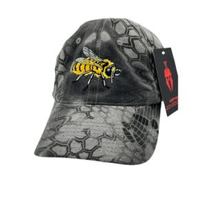Puede incluir: Gorra de béisbol de camuflaje gris y negro con una abeja bordada amarilla y negra en la parte delantera. La gorra tiene una visera curva y una etiqueta con un logotipo rojo.