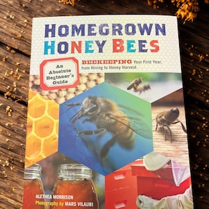 Puede incluir: Un libro titulado "Homegrown Honey Bees" con el subtítulo "Beekeeping Your First Year, from Hiving to Honey Harvest." La portada presenta una abeja, un panal, tarros de miel y un apicultor. La autora es Alethea Morrison.