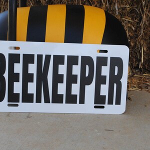 Puede incluir: Un letrero blanco con texto negro que dice "BEEKEEPER". El letrero est&aacute; apoyado contra un objeto con forma de abeja negro y amarillo.