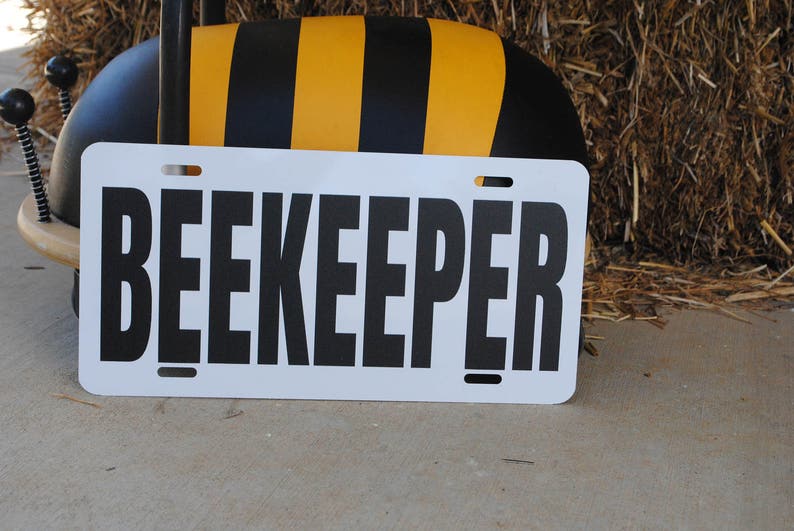 Puede incluir: Un letrero blanco con texto negro que dice "BEEKEEPER". El letrero est&aacute; apoyado contra un objeto con forma de abeja negro y amarillo.