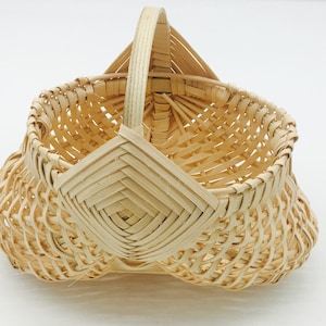 Peut inclure: Un petit panier tissé avec une poignée. Le panier est fait de bois clair et naturel et présente un motif en forme de losange sur le devant. La poignée est arquée et fixée aux côtés du panier.