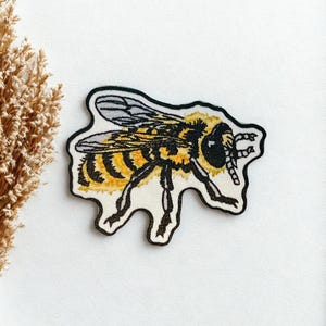 May include: Embroidered bee patch with yellow and black details, outlined in black. The patch features a detailed design of a bee with visible wings and legs. The patch is on a white background, with a dried plant stem on the left.