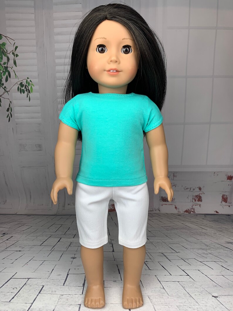 May include: A doll wearing a turquoise short-sleeved shirt and white shorts. The doll has long black hair and brown eyes.