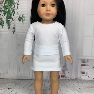 May include: A white long-sleeved shirt and a white skirt outfit for a doll. The doll has long black hair and brown eyes.