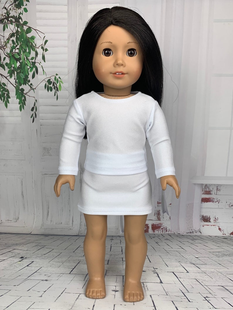 May include: A white long-sleeved shirt and a white skirt outfit for a doll. The doll has long black hair and brown eyes.