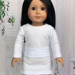 May include: A white long-sleeved shirt and skirt outfit for a doll. The doll is wearing a white shirt with long sleeves and a white skirt. The doll has long black hair and brown eyes.