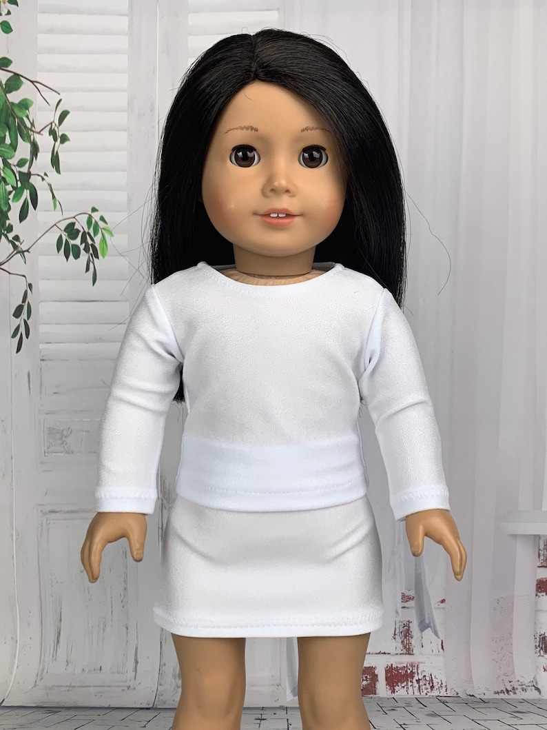 May include: A white long-sleeved shirt and skirt outfit for a doll. The doll is wearing a white shirt with long sleeves and a white skirt. The doll has long black hair and brown eyes.