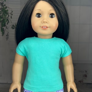 May include: A doll wearing a turquoise t-shirt and purple floral print shorts.