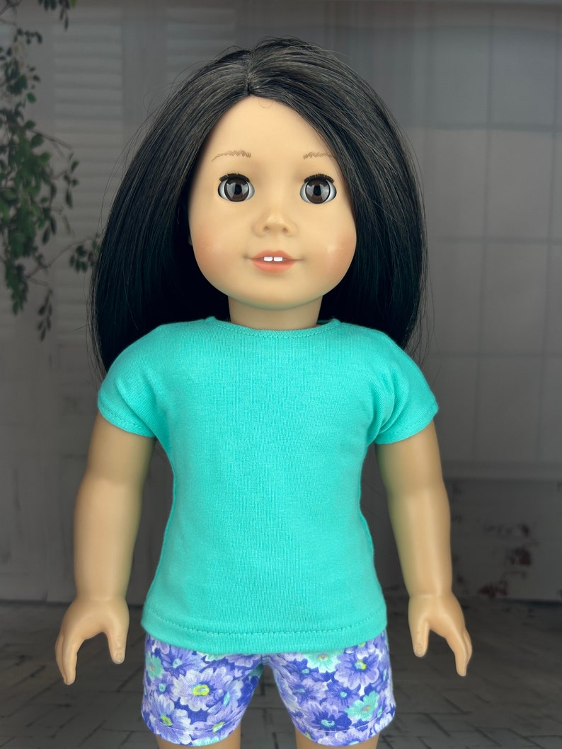 May include: A doll wearing a turquoise t-shirt and purple floral print shorts.