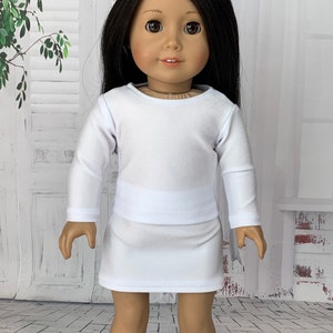 May include: A white long-sleeved shirt and skirt set for a doll. The outfit is made of a soft, stretchy fabric and has a simple, classic design.