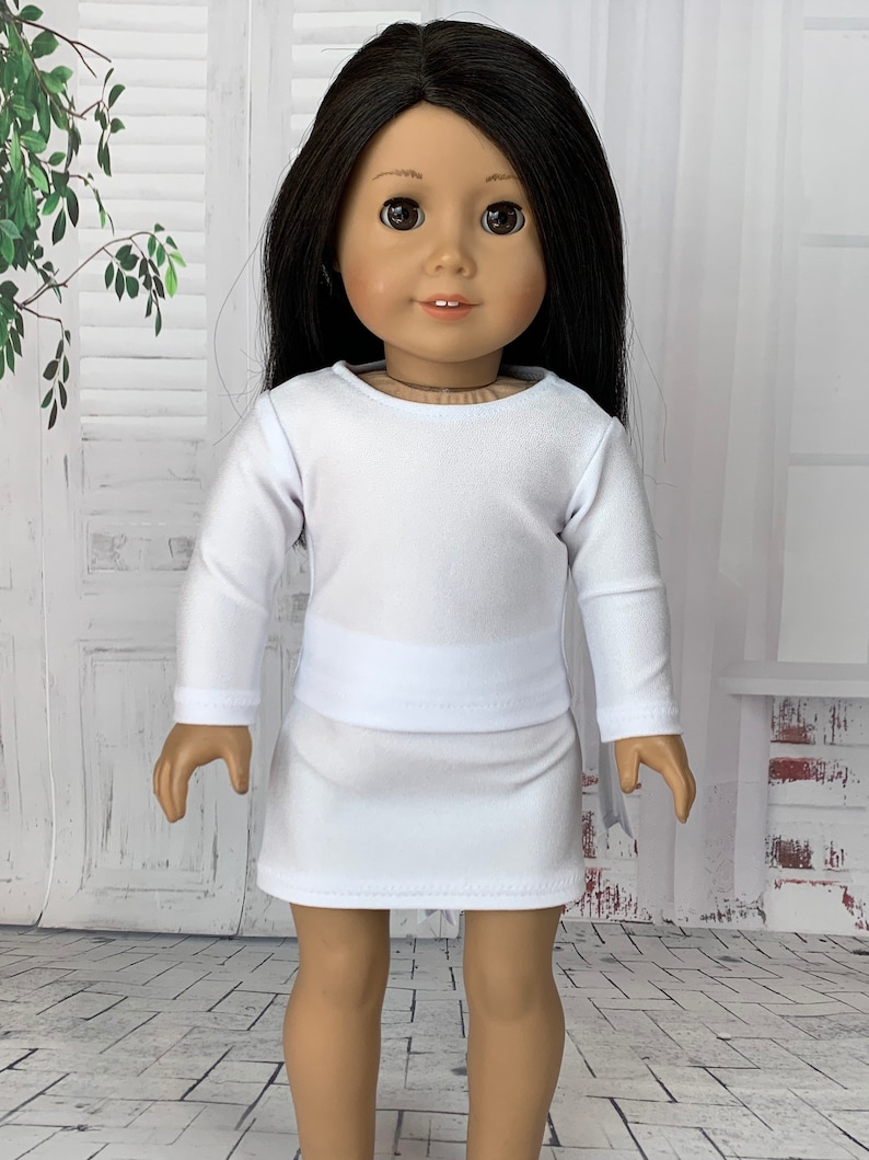 May include: A white long-sleeved shirt and skirt set for a doll. The outfit is made of a soft, stretchy fabric and has a simple, classic design.